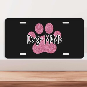 May include: Black license plate with a pink glitter paw print and the text "Dog MiMi". The plate is made of aluminum and measures 12 inches by 6 inches. It has four holes for attaching to a car.