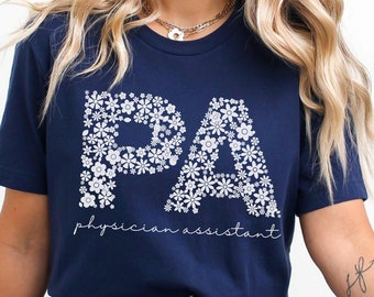 May include: Navy blue t-shirt with a white floral design spelling out "PA" and the text "physician assistant".