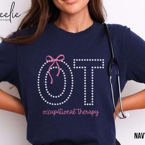 May include: Navy blue t-shirt with the letters "OT" in white dotted font, a pink bow, and the words "occupational therapy" in pink. A stethoscope hangs on the side. The shirt is labeled "NAVY".