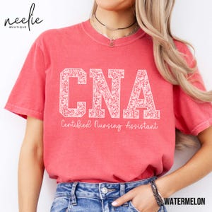 May include: A red t-shirt with a white floral design that spells out "CNA" and the text "Certified Nursing Assistant".