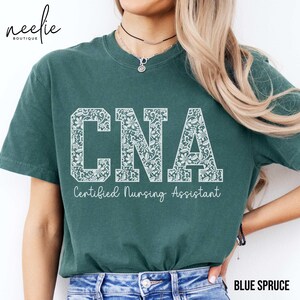 May include: A teal blue t-shirt with a white lace design that spells out "CNA" and the text "Certified Nursing Assistant" underneath.
