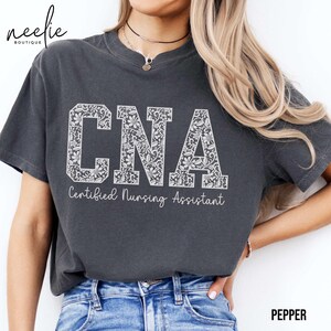 May include: A dark gray t-shirt with a white lace design spelling out "CNA" and the text "Certified Nursing Assistant" underneath.