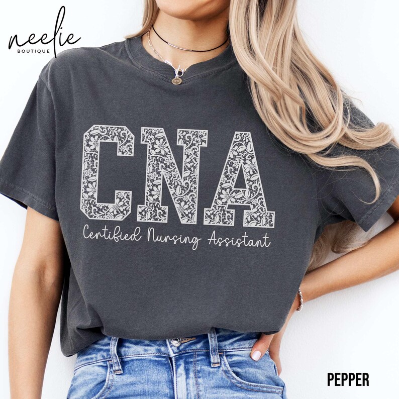 May include: A dark gray t-shirt with a white lace design spelling out "CNA" and the text "Certified Nursing Assistant" underneath.