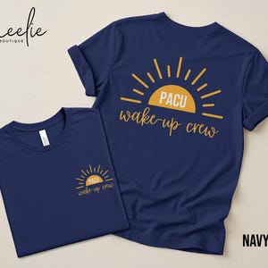 May include: Two navy blue t-shirts with the text "PACU wake-up crew" printed in gold. One shirt has the design on the front, the other on the back. The shirts are displayed on a light beige surface.