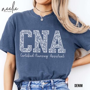 May include: A blue t-shirt with the words "CNA Certified Nursing Assistant" printed in white with a floral design. The shirt is being worn by a person with long blonde hair.