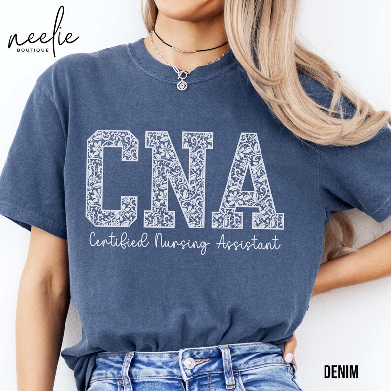 May include: A blue t-shirt with the words "CNA Certified Nursing Assistant" printed in white with a floral design. The shirt is being worn by a person with long blonde hair.