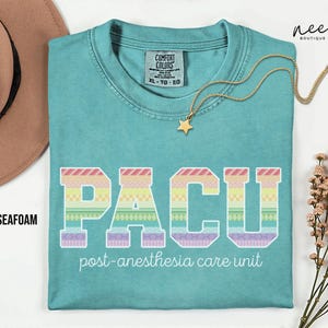 May include: Seafoam green t-shirt with the text "PACU" in a colorful, patterned design. Below the letters, the words "post-anesthesia care unit" are printed. A gold star pendant necklace is draped across the shirt.