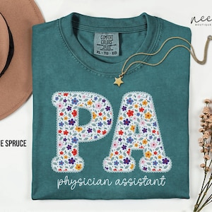 May include: Blue spruce t-shirt with the letters "PA" in a floral design, with the words "physician assistant" below. A gold necklace with a star pendant is on the shirt.