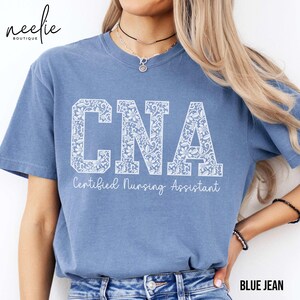May include: A blue jean t-shirt with a white lace design that says "CNA Certified Nursing Assistant".