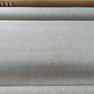 May include: A roll of light gray fabric featuring a visible woven texture with subtle vertical variations in tone. The material is tightly wound, displaying a consistent, neutral color palette across the entire surface of the textile roll.