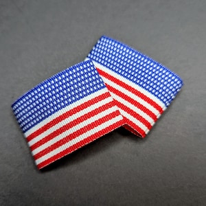 May include: Two fabric ribbons, each featuring a design reminiscent of the American flag. The ribbons display a blue field of white stars and red and white stripes, set against a dark gray background.