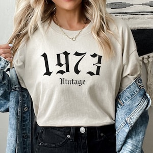 Op de afbeelding: Beige shirt met lange mouwen met het jaar "1973" in een gotisch lettertype en het woord "Vintage" eronder. Het shirt wordt gedragen met een spijkerjasje en een zwarte jeans. Een gouden ketting is zichtbaar.