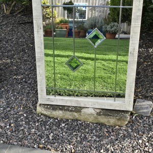 May include: A rectangular outdoor mirror with a distressed white frame. The mirror has a grid pattern with two hanging green diamond-shaped glass accents. The mirror reflects a green lawn and potted plants.