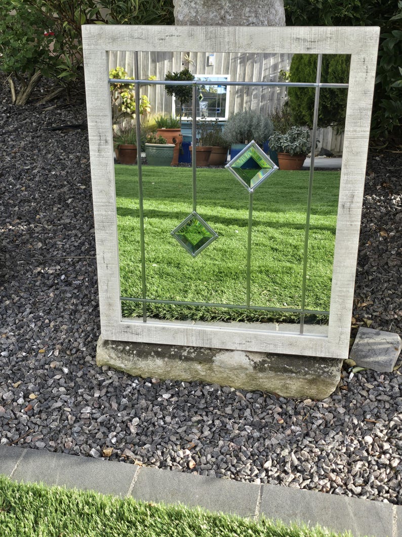May include: A rectangular outdoor mirror with a distressed white frame. The mirror has a grid pattern with two hanging green diamond-shaped glass accents. The mirror reflects a green lawn and potted plants.