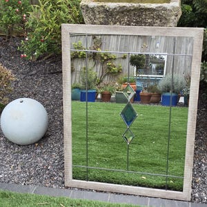 May include: A rectangular mirror with a light-colored wooden frame. The mirror has a decorative metal grid with two diamond-shaped glass accents. The mirror is placed on a green lawn, reflecting the surrounding garden.