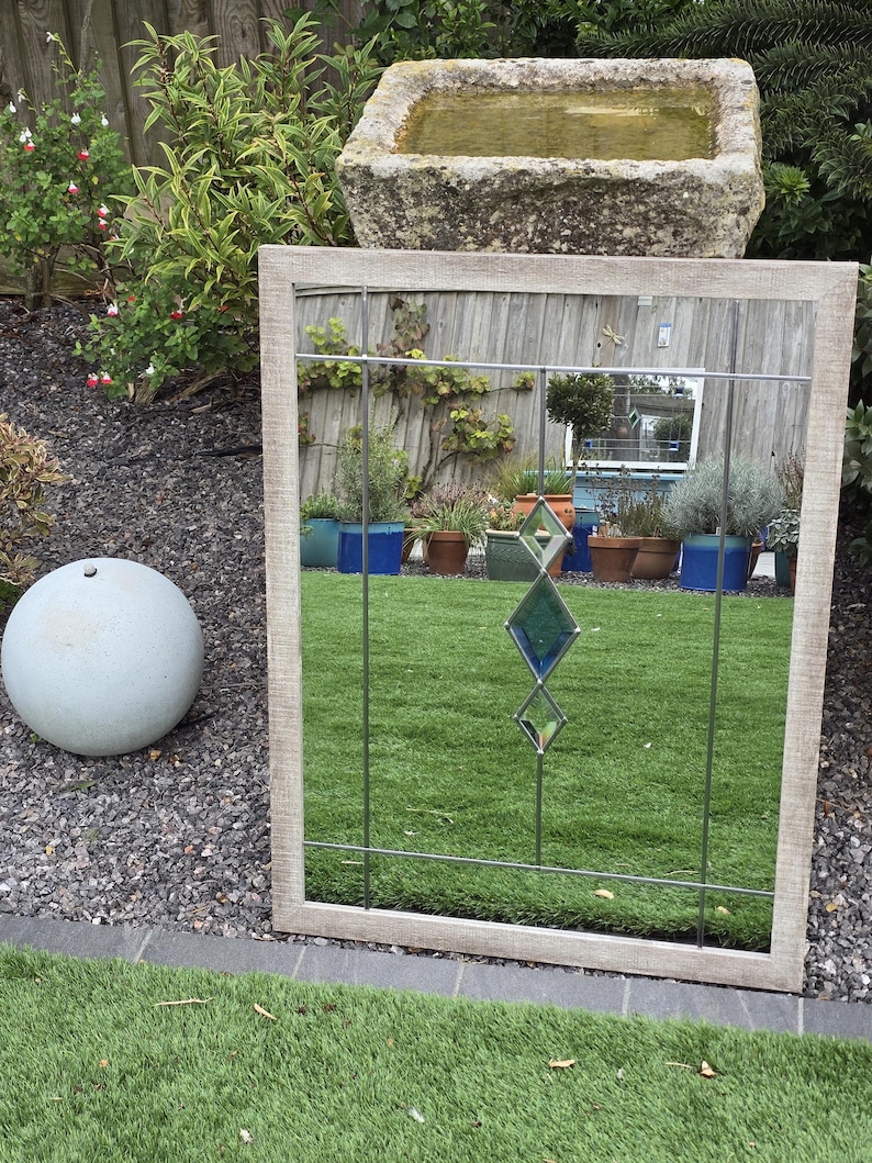 May include: A rectangular mirror with a light-colored wooden frame. The mirror has a decorative metal grid with two diamond-shaped glass accents. The mirror is placed on a green lawn, reflecting the surrounding garden.