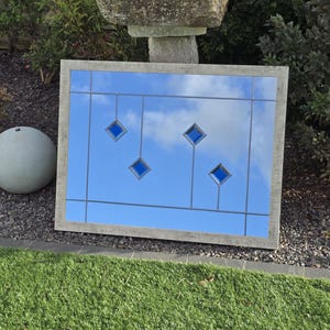 May include: A rectangular mirror with a light gray wooden frame. The mirror reflects a blue sky with clouds and features four blue diamond-shaped accents. A gray sphere is to the left of the mirror, and green grass is in the foreground.