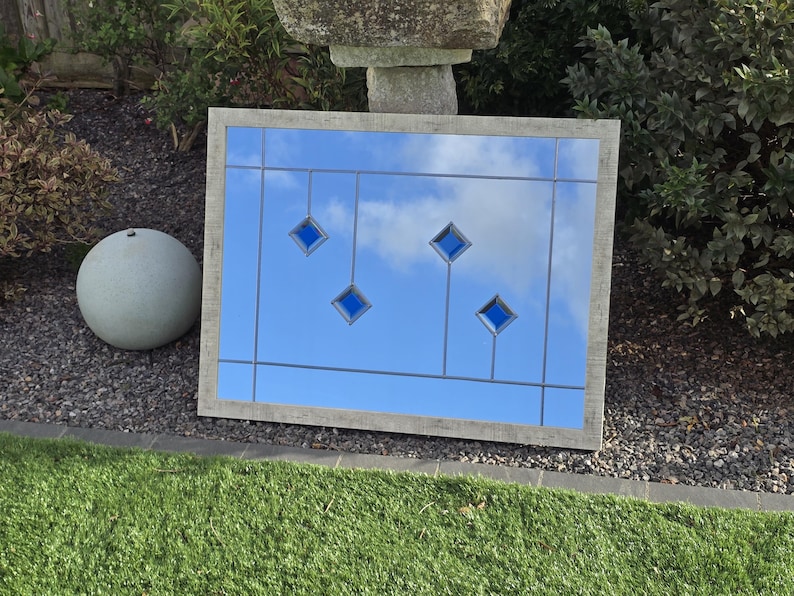 May include: A rectangular mirror with a light gray wooden frame. The mirror reflects a blue sky with clouds and features four blue diamond-shaped accents. A gray sphere is to the left of the mirror, and green grass is in the foreground.