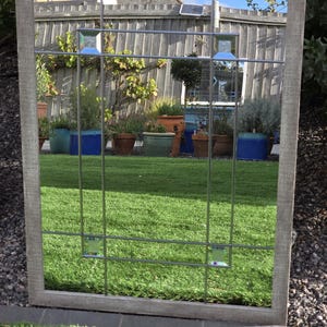 May include: A rectangular mirror with a gray wooden frame and a grid of metal bars. The mirror reflects a backyard scene with green grass, potted plants, and a wooden fence. Decorative glass squares are set into the metal grid.