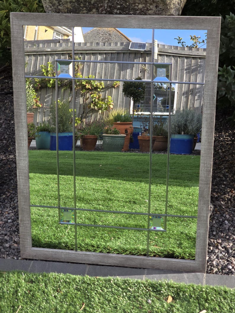 May include: A rectangular mirror with a gray wooden frame and a grid of metal bars. The mirror reflects a backyard scene with green grass, potted plants, and a wooden fence. Decorative glass squares are set into the metal grid.