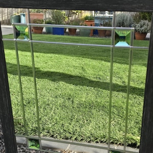 May include: A decorative window with a black wooden frame and a grid pattern. The window features clear glass panes with green square accents. The background shows a green lawn and potted plants.