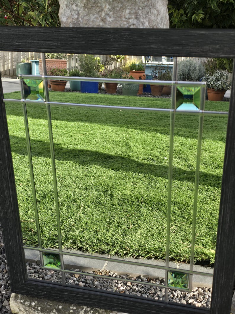 May include: A decorative window with a black wooden frame and a grid pattern. The window features clear glass panes with green square accents. The background shows a green lawn and potted plants.