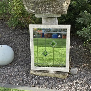 May include: A rectangular outdoor mirror with a weathered white frame. The mirror features a grid pattern with three green diamond-shaped accents. The mirror is propped up on a stone base in a garden setting.