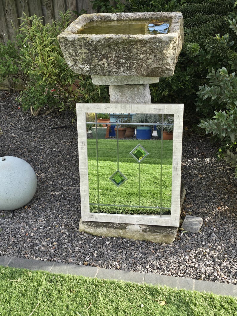 May include: A rectangular outdoor mirror with a weathered white frame. The mirror features a grid pattern with three green diamond-shaped accents. The mirror is propped up on a stone base in a garden setting.
