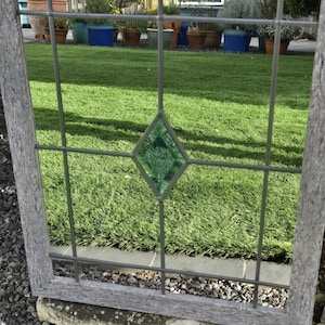 May include: A weathered wooden window frame with a grid pattern and a central green diamond-shaped glass insert. The window reflects a vibrant green lawn and potted plants, creating a rustic, outdoor aesthetic.