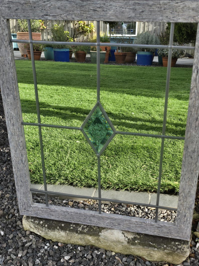 May include: A weathered wooden window frame with a grid pattern and a central green diamond-shaped glass insert. The window reflects a vibrant green lawn and potted plants, creating a rustic, outdoor aesthetic.