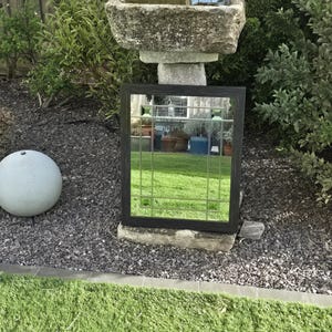 May include: A black framed mirror with a grid pattern, resting on a stone base in a garden setting. The mirror reflects the surrounding greenery and sky. A stone bird bath is visible in the background.