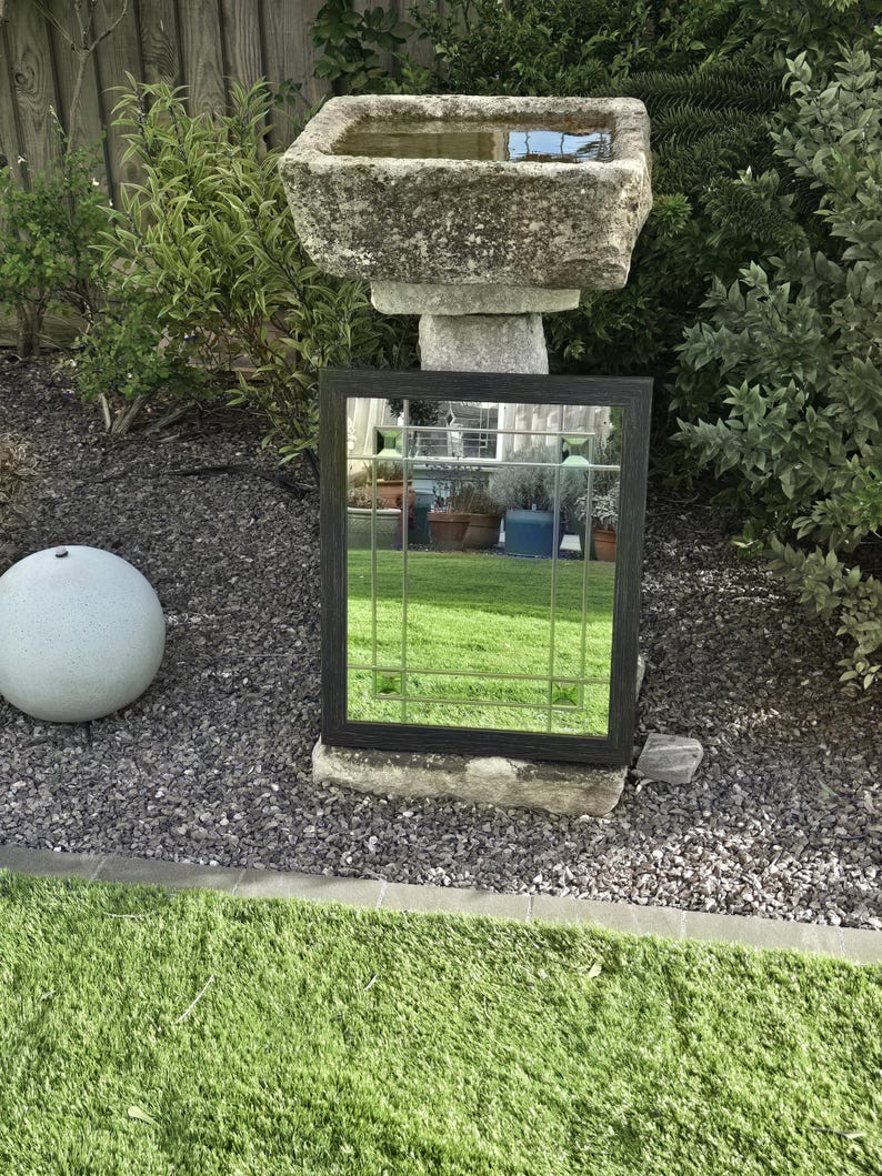 May include: A black framed mirror with a grid pattern, resting on a stone base in a garden setting. The mirror reflects the surrounding greenery and sky. A stone bird bath is visible in the background.