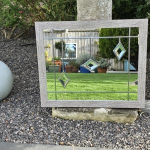 May include: A rectangular mirror with a weathered gray wooden frame. The mirror features a grid of thin metal lines and three hanging diamond-shaped glass accents. The mirror reflects a green lawn, plants, and a building.