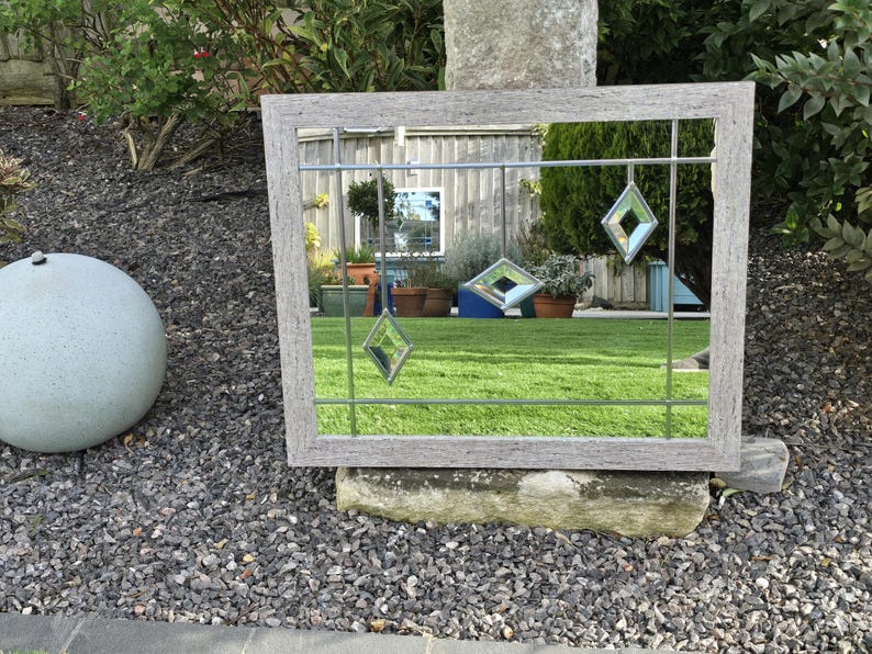 May include: A rectangular mirror with a weathered gray wooden frame. The mirror features a grid of thin metal lines and three hanging diamond-shaped glass accents. The mirror reflects a green lawn, plants, and a building.