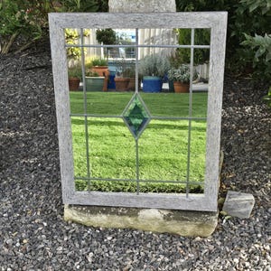 May include: A rectangular mirror with a gray wooden frame and a grid pattern. The mirror has a green diamond-shaped accent in the center. The mirror is placed outdoors, reflecting a garden scene with green grass and potted plants.