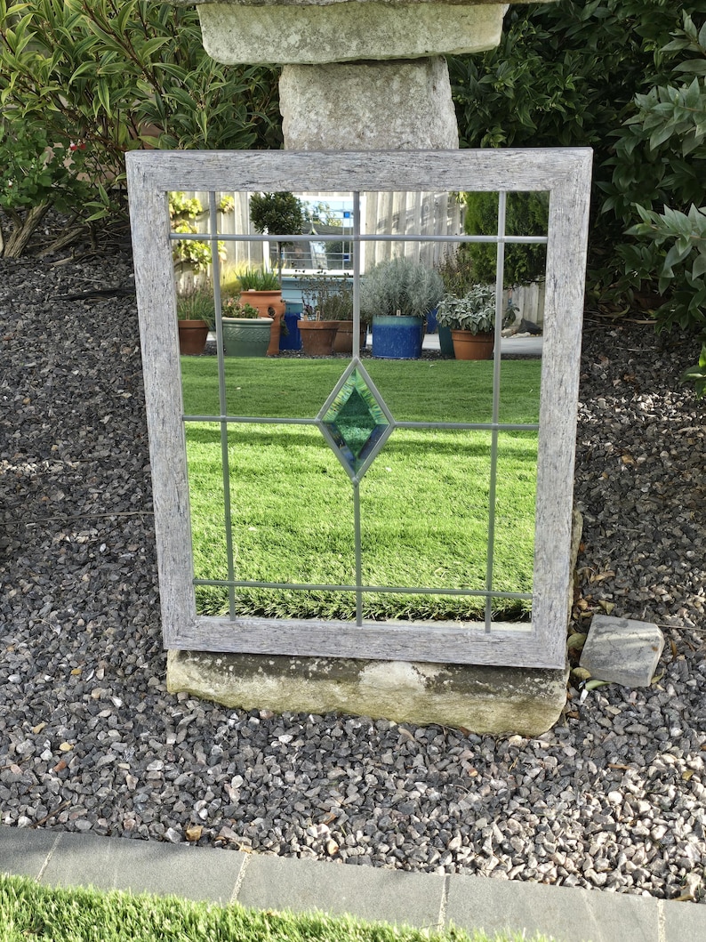 May include: A rectangular mirror with a gray wooden frame and a grid pattern. The mirror has a green diamond-shaped accent in the center. The mirror is placed outdoors, reflecting a garden scene with green grass and potted plants.