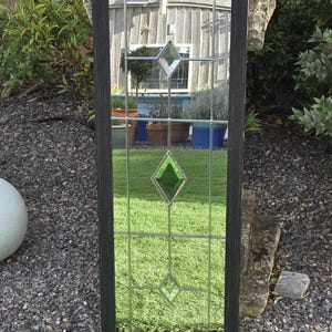 K&ouml;nnte beinhalten: Rechteckiger Spiegel mit dunklem Rahmen und Buntglasdesign. Das Glas hat ein Gittermuster mit rautenf&ouml;rmigen Akzenten in Klar und Gr&uuml;n. Der Spiegel befindet sich im Freien und spiegelt eine Gartenszene wider.
