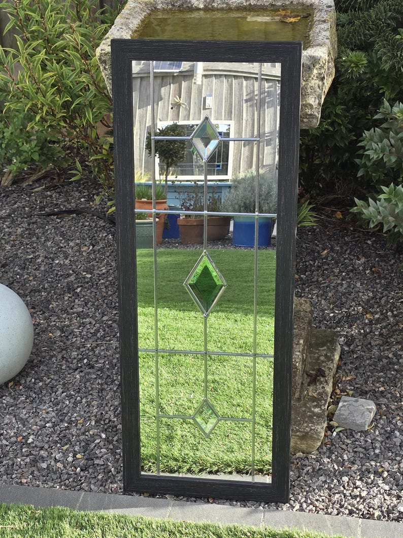 K&ouml;nnte beinhalten: Rechteckiger Spiegel mit dunklem Rahmen und Buntglasdesign. Das Glas hat ein Gittermuster mit rautenf&ouml;rmigen Akzenten in Klar und Gr&uuml;n. Der Spiegel befindet sich im Freien und spiegelt eine Gartenszene wider.