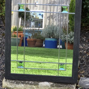 May include: Rectangular mirror with a dark gray wooden frame. The mirror features a grid design with small, square, green glass accents. The reflection shows a garden with potted plants and green grass.