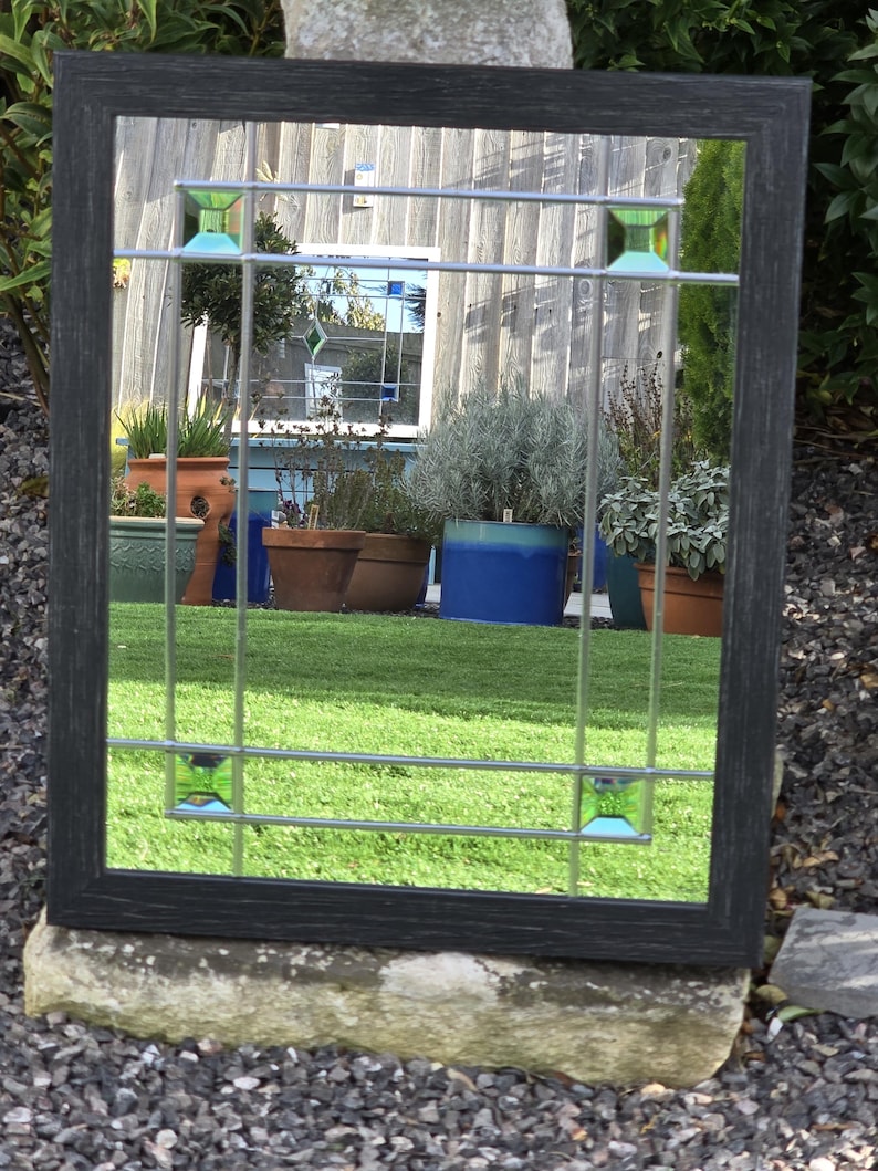 May include: Rectangular mirror with a dark gray wooden frame. The mirror features a grid design with small, square, green glass accents. The reflection shows a garden with potted plants and green grass.