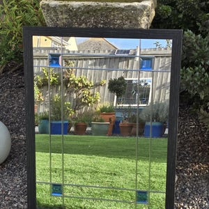 May include: A rectangular mirror with a dark wood frame and a grid pattern. The mirror has blue and teal accents. The reflection shows a green lawn, plants, and a stone water trough. The mirror is outdoors.