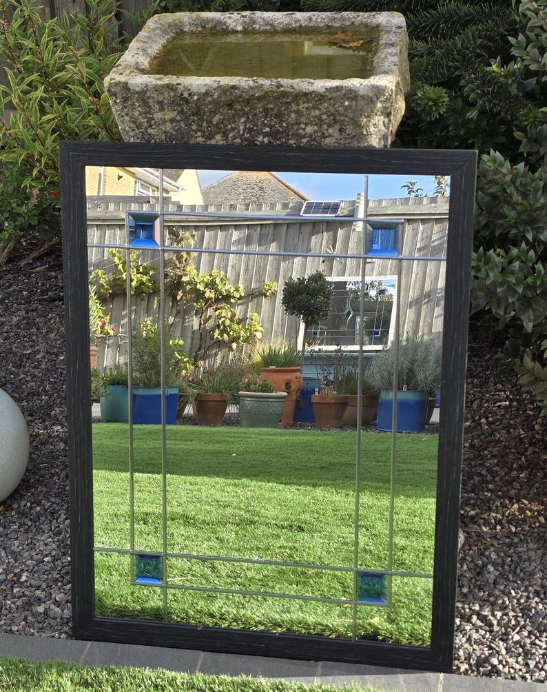 May include: A rectangular mirror with a dark wood frame and a grid pattern. The mirror has blue and teal accents. The reflection shows a green lawn, plants, and a stone water trough. The mirror is outdoors.