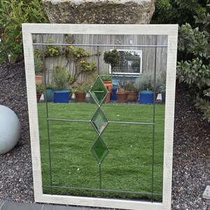 May include: A rectangular mirror with a white, textured frame. The mirror features a grid pattern with three green diamond-shaped glass inserts. The reflection shows a garden scene with plants and a stone water trough.