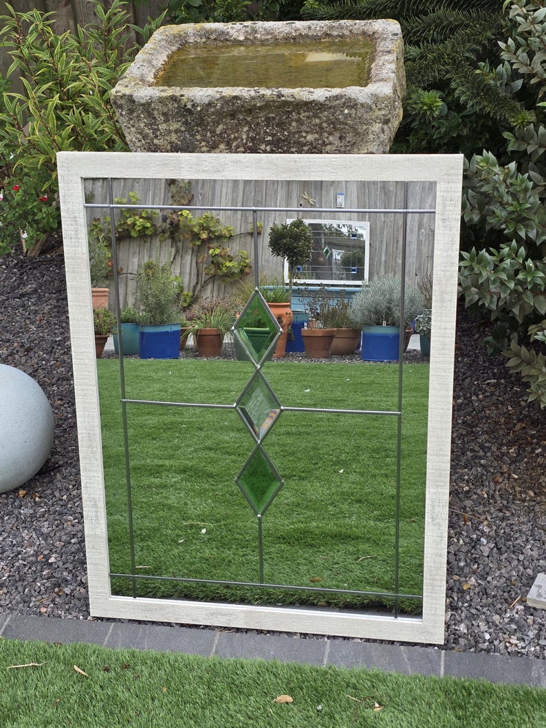 May include: A rectangular mirror with a white, textured frame. The mirror features a grid pattern with three green diamond-shaped glass inserts. The reflection shows a garden scene with plants and a stone water trough.
