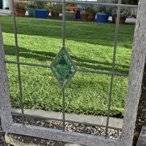 May include: A weathered, gray wooden window frame with a grid of metal bars. A diamond-shaped, green stained-glass element is centered in the frame. The window overlooks a green lawn and potted plants.