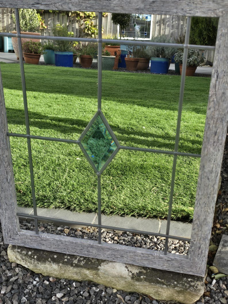 May include: A weathered, gray wooden window frame with a grid of metal bars. A diamond-shaped, green stained-glass element is centered in the frame. The window overlooks a green lawn and potted plants.