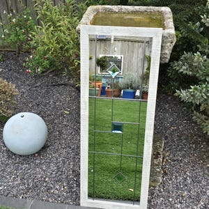 May include: A white-framed rectangular mirror with a grid design and a central blue square and diamond. The mirror reflects a garden scene with plants and a stone birdbath. The frame has a distressed finish. The mirror is outdoors.