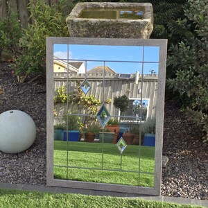 May include: A rectangular mirror with a gray wooden frame and a grid pattern. The mirror reflects a garden scene with green grass, plants, and a stone water feature. Decorative diamond-shaped glass accents are visible.