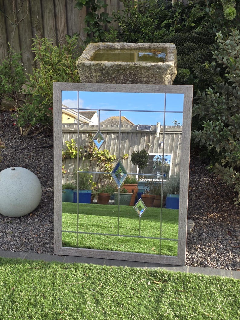 May include: A rectangular mirror with a gray wooden frame and a grid pattern. The mirror reflects a garden scene with green grass, plants, and a stone water feature. Decorative diamond-shaped glass accents are visible.