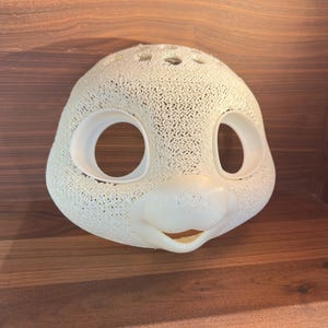 May include: A cream-colored, 3D-printed mask shaped like a cartoon character's face. The mask has large, oval eye openings and a small, smiling mouth. The surface has a textured, porous pattern. The mask is displayed on a wooden surface.